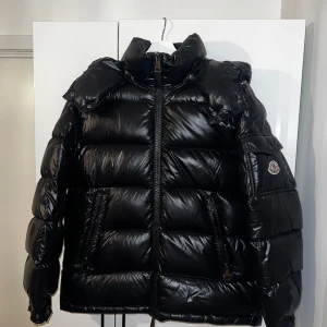 Svart Moncler pufferjacka dam - Säljer en svart pufferjacka från Moncler med glansig finish, hög krage och huva. Jackan har dragkedja framtill, fickor med dragkedja och en Moncler-logga på ärmen. Modellen är MAIRE, storlek 1 vilket motsvarar en EU S. Kvitto finns även tillgängligt. Finns en liten skrapa på insidan av jackan, bara nån millimeter som knappt syns. 