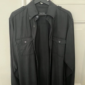 Tom Ford skjorta - Svart overshirt från Tom Ford med klassisk krage, axelklaffar och två bröstfickor med knappar. Skjortan har lång ärm och är tillverkad i mjuk bomull. Perfekt för en clean och stilren look.