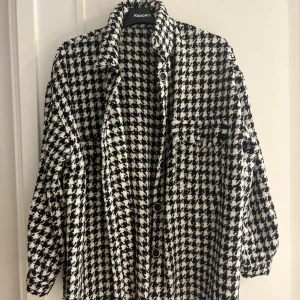 Overshirt i svartvitt hundtandsmönster  - Snygg overshirt  i svart och vitt hundtandsmönster. Jackan har stora knappar framtill, bröstficka med fransad kant och en lite ruffig känsla. Perfekt för lager-på-lager och ger en cool vibe till din outfit.