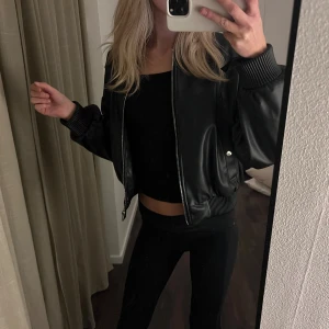 Svart bomberjacka i skinn från Zara - Snygg svart bomberjacka från Zara i skinnimitation med ribbade muddar och krage. Jackan har dragkedja framtill och två fickor med knapp.