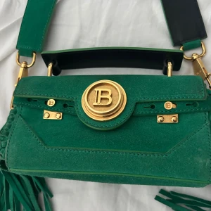 Grön axelväska från Balmain i mocka - Snygg grön axelväska från Balmain i mjuk mocka med bred axelrem och guldiga metalldetaljer. Väskan har fransar på sidorna, ett stort guldfärgat B-spänne framtill och flera dekorativa nitar. Insidan är ljus och har ett fack för småsaker.