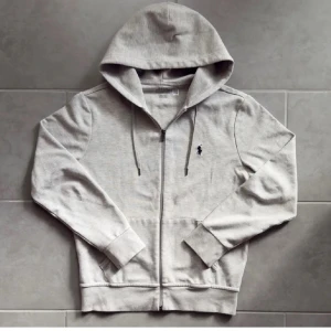 Grå hoodie från Polo Ralph Lauren - Snygg ljusgrå hoodie från Polo Ralph Lauren med dragkedja framtill och klassisk broderad logga på bröstet. Tröjan har huva med snörning, ribbade muddar och kängurufickor. Perfekt för chill dagar eller när du vill ha en clean look.