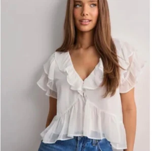 Vit volangblus med v-ringning - Supersöt vit blus från Nelly med v-ringning och volangdetaljer längs ärmar och nederkant. Blusen är gjord i ett tunt, luftigt material och har korta ärmar. Perfekt att styla med jeans eller kjol för en fräsch look.