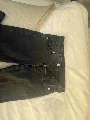 Svarta bootcut Low jeans - Säljer ett par svarta bootcut jeans med klassisk femficksdesign och knappgylf. Jeansen har en snygg utsvängd passform och är tillverkade i ett mjukt bomullsmaterial med lite stretch. Perfekta för dig som gillar en retroinspirerad look.