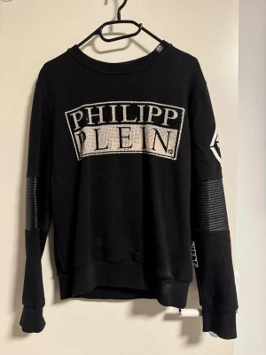 Svart sweatshirt från Philipp Plein - Säljer en svart sweatshirt från Philipp Plein med stort logotryck framtill och coola detaljer på ärmarna. Tröjan har rund halsringning och ribbade muddar. Perfekt för dig som gillar streetwear och vill sticka ut lite extra. Slitage och saknade stenar 