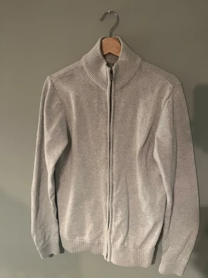 Ljusgrå kofta med dragkedja - Säljer en stilren ljusgrå kofta från Dobber med hel dragkedja framtill och ribbad hög krage. Tröjan har långa ärmar och ribbade muddar vid ärmslut och nederkant. Perfekt för lager på lager under kyliga dagar.