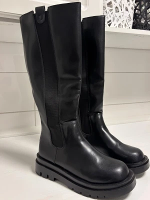 Svarta höga boots med dragkedja - Säljer ett par svarta höga boots i skinnimitation med chunky sula och dragkedja baktill. Har använt de 1 gång! Den är märkt med storlek 43, men är storlek 41.5. De är breda vid vaderna.