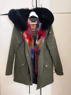 Moose Knuckles parkas med färgglad päls - Säljer en grön parkas från Moose Knuckles med stor svart pälskantad huva och färgglad pälsdetalj på insidan. Jackan har guldfärgade metalldetaljer på ärmen, dragkedja och tryckknappar. Insidan är quiltad och fodrad för extra värme.