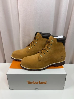 Timberland boots i mocka, beige - Klassiska Timberland boots i beige mocka med bruna detaljer och kraftig gummisula. Skorna har snörning med guldfärgade öljetter och vadderad krage för extra komfort. Perfekta för höst och vinter, ikonisk streetwear-vibe.