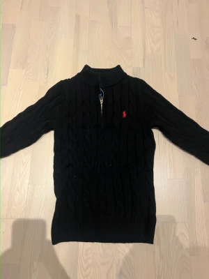 Svart stickad tröja Polo Ralph Lauren - Kabelstickad svart tröja från Polo Ralph Lauren med halv dragkedja och röd broderad logga på bröstet. Tröjan har hög krage och långa ärmar, perfekt för kyligare dagar. Klassisk och tidlös design som funkar till allt.