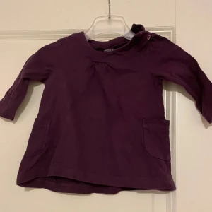 Mörklila babyblus med fickor - Supersöt mörklila blus för baby med trekvartsärm och två praktiska fickor framtill. Blusen har en knappdetalj vid axeln för enkel påklädning och är gjord i mjuk bomull som känns skön mot huden.