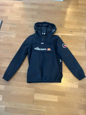 Svart vindjacka från Ellesse - Snygg svart vindjacka från Ellesse med stor ficka över magen. Hyffsat tunn vindkacka, bra skick men finns mindre skavanker i färgen på vissa ställen.