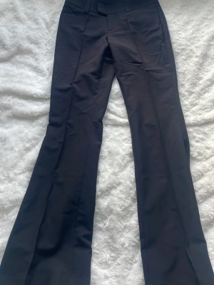 Svarta bootcut kostymbyxor - Snygga svarta kostymbyxor med bootcut passform och hög midja. Byxorna har pressveck framtill, bälteshällor och två fickor med lock bak. Perfekta för en clean och stilren look. Materialet känns mjukt och följsamt.