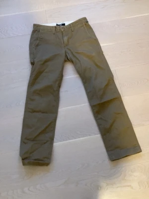 Beiga chinos från Vans - Säljer ett par klassiska beiga byxor  från Vans. Byxorna har raka ben, normal passform och bälteshällor. Tillverkade i mjuk bomull med två fickor fram och två bakfickor. Perfekta för en clean och avslappnad stil.