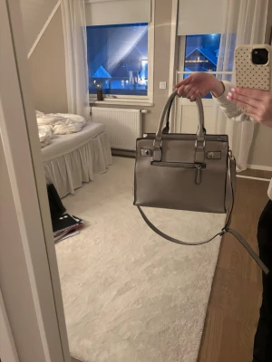 Grå handväska med axelrem - Snygg grå handväska med både handtag och justerbar axelrem. Väskan har en stilren, fyrkantig form och är tillverkad i skinnimitation. Utsidan har en dragkedja framtill och insidan är uppdelad i flera fack, varav ett med dragkedja. Perfekt för dig som gillar organiserad förvaring.