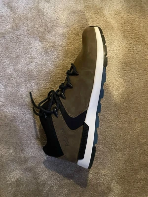 Timberland boots i mocka och textil - Säljer ett par snygga Timberland boots i olivbrun mocka och svart textil med vita och svarta detaljer. Skorna har grov sula, snörning och en sportig, modern look. Perfekta för dig som vill ha varma men snygga skor