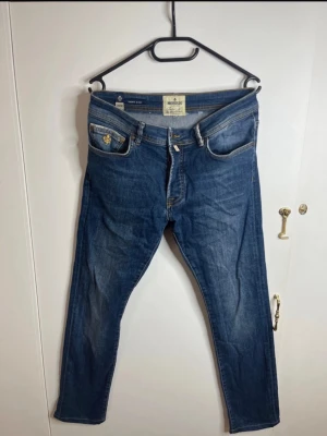 Morris Triumph jeans blå 31/32 - Snygga Morris Triumph jeans i klassisk blå tvätt med gul broderad detalj på fickan och coola bakfickor med zigzag-söm. Modellen har normal passform och raka ben, tillverkade i slitstarkt bomullsmaterial. Perfekt för dig som gillar stilrena jeans med lite extra detaljer.