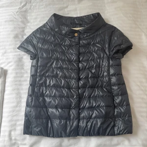 Svart kortärmad väst/cape från Herno - Säljer en svart, kortärmad pufferjacka från Herno med hög krage och tryckknappar framtill. Storlek 40, passar en XS & S, Nypris ca 4700kr🤍pris kan diskuteras.