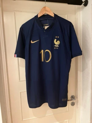 France home kit 22 X Mbappe 10 - Säljer en officiell fransk landslagströja från Nike med Mbappé och nummer 10 i guld. Tröjan är marinblå med kort ärm, Dri-FIT-material och snygga gulddetaljer. Perfekt för fotbollsfans som vill sticka ut på läktaren eller på planen.