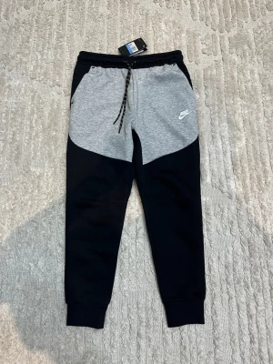 Nike mjukisbyxor svart/grå  - Nike mjukisbyxor i storlek M med snygg tvåfärgad design i svart och ljusgrått. Byxorna har snörning i midjan, ribbade muddar och logga på vänster lår. Tillverkade i mjukt bomullsmaterial, perfekta för chill eller träning. Byxorna är i storlek M men skulle säga är mer som XS och S 