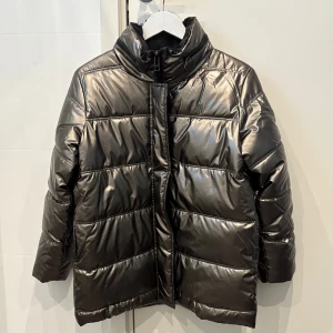 Metallic silver pufferjacka Zadig&Voltaire - Säljer en metallic silver pufferjacka från Zadig&Voltaire med hög krage och dragkedja framtill. Jackan har en glansig finish, raka ärmar och quiltad design som ger en riktigt cool vibe. Perfekt för dig som vill sticka ut under kalla dagar. Dold luva i kragen. Varm och skön och går ner över baken. Helt oanvänd = nyskick! Hel och ren och inget är slitet. Storlek 14 år eller XS.