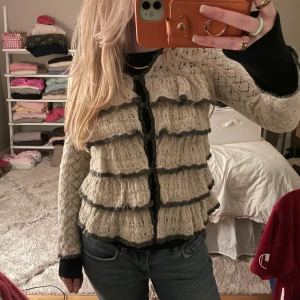 Beige kofta från Odd Molly med volanger - Supersöt beige kofta från Odd Molly med virkade detaljer och volanger i flera lager. Koftan har mörkgrå kanter, v-ringning och knappar framtill. Långärmad och stickad i ett luftigt mönster som ger en bohemisk vibe.