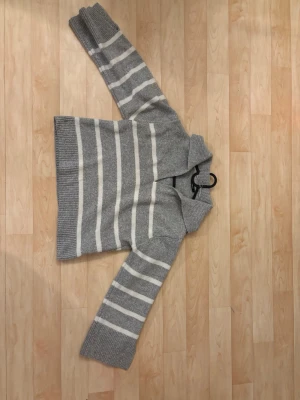 Gråvit randig stickad tröja Gina Tricot - Mysig stickad tröja från Gina Tricot i storlek M. Tröjan är grå med vita horisontella ränder, har en bred krage och långa ärmar med ribbade muddar. Materialet är en blandning av polyester, akryl, polyamid och ull. Perfekt för kyliga dagar.