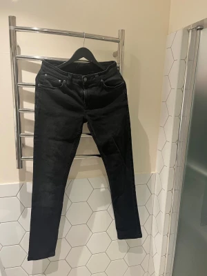 Svarta jeans från Nudie Jeans - Säljer ett par svarta jeans från Nudie Jeans med  snygga detaljer på bakfickorna. Jeansen har en mörk tvätt och är tillverkade i stretchig bomull för skön passform. Perfekt för dig som gillar stilrena jeans. Bredd 30 längd 34 