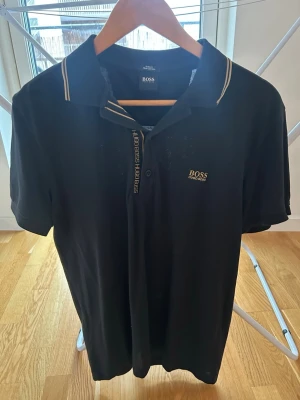 Svart piké från Hugo Boss - Säljer en svart pikétröja från Hugo Boss i storlek L. Tröjan har guldfärgade detaljer på kragen och logga på bröstet. Slim fit-modell i mjuk pima bomull med knappslå och klassisk krage. Perfekt för en clean och stilren look.
