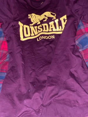 Vinröd Lonsdale t-shirt i bomull - Säljer en vinröd t-shirt från Lonsdale London med stort gult tryck framtill. Klassisk rund halsringning och korta ärmar. Skön och mjuk i bomull, perfekt till en avslappnad stil. Passformen är normal och materialet känns lätt och bekvämt. Använd 2 gånger 
