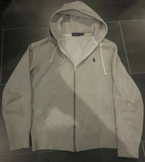 Grå hoodie från Polo Ralph Lauren - Snygg ljusgrå hoodie från Polo Ralph Lauren med dragkedja och huva. Klassisk design med två fickor fram, vita snören och broderad logga på bröstet. Tillverkad i mjuk bomullsmix, perfekt för chill dagar eller när du vill ha en clean look.