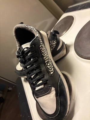 Svarta och vita sneakers från Hugo - Snygga sneakers från Hugo med svart och vit färg, coolt grafiskt mönster på sidorna och svarta snören. Skorna har rund tå, platt sula och detaljer i både skinn och syntetmaterial. Perfekta för dig som gillar streetstyle och vill sticka ut med ett unikt mönster.