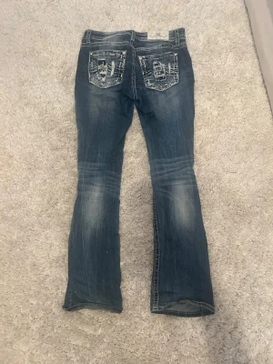Miss Me blå bootcut jeans med detaljer - Säljer ett par blå jeans från Miss Me i bootcut-modell. Jeansen har slitna detaljer och dekorerade bakfickor med broderier och nitar. Tydliga kontrastsömmar och snygg tvätt ger dem en unik look. Perfekta för dig som vill sticka ut lite extra! 
