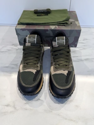 Valentino Garavani Rockrunner  - Snygga Valentino Garavani Rockrunner sneakers i camouflage med gröna, svarta och beige toner. Skorna har ovandel i mesh, mocka och skinn, samt coola nitar på hälen. Klassisk snörning och platt sula gör dem både trendiga och bekväma.
