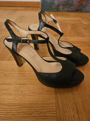 Svarta klackade sandaler i skinn - Snygga svarta sandaler med hög klack och öppen tå. Skorna har T-rem med spänne och plattform framtill för extra komfort. Tillverkade i skinn med beige innersula. Perfekta för dig som vill ha en stilren och klassisk look.
