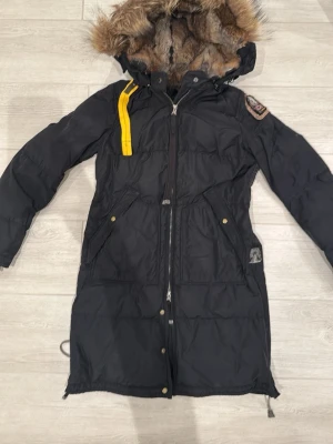 Svart Parajumpers parkas med fuskpäls - Lång svart parkas från Parajumpers med stor huva fodrad i mjuk fuskpäls. Jackan har en gul detalj vid axeln, patch på ärmen och dragkedja framtill. Två stora fickor med knappar och justerbar nederkant. Perfekt för kalla dagar.