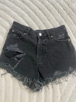 Svarta denimshorts med slitningar - Säljer ett par svarta högmidjade jeansshorts från h&m med råa, fransiga kanter och slitna detaljer framtill. Klassisk femficksmodell med knapp. Perfekta för en avslappnad och edgy stil. Pris går att diskutera.