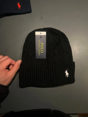 Svart mössa från Polo Ralph Lauren - Svart ribbstickad mössa från Polo Ralph Lauren med klassisk vit broderad logga framtill. Tillverkad i mjuk ull som håller dig varm och snygg under kyliga dagar. Enkel och stilren design som passar till det mesta.