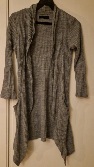 Grå ribbad kofta från Vero Moda - Säljer en lång, gråmelerad ribbad kofta från Vero Moda i storlek M. Koftan har öppen front, långa ärmar och två fickor framtill. Materialet är mjukt och stretchigt. Hund finns i hemmet. Kolla in mina andra annonser för samfrakt! 