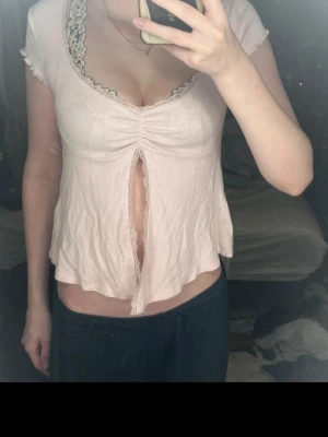 Ljusrosa blus med spets och slits - Supersöt ljusrosa blus med korta ärmar och djup v-ringning dekorerad med spets. Toppen har en unik slits framtill och rynkdetalj vid bysten. Materialet känns mjukt och lätt, perfekt för en trendig look.