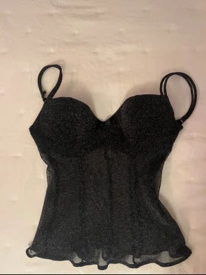 Glittrig svart korsettopp Intimissimi - Supersnygg svart korsettopp från Intimissimi med glittrigt tyg och tunna dubbla axelband. Toppen har lätt vadderade kupor och en liten rosett framtill. Materialet är genomskinligt och ger en lyxig känsla, perfekt för att sticka ut.