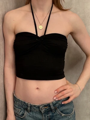 Svart bandeau tubtopp från H&M - Säljer en svart bandeau tubtopp från H&M i storlek XS. Toppen har halterneck-band med pärldetaljer och snygg twistad front. Materialet är lätt ribbat och stretchigt, perfekt till jeans eller kjol. Knytbandet i nacken ger en cool look.