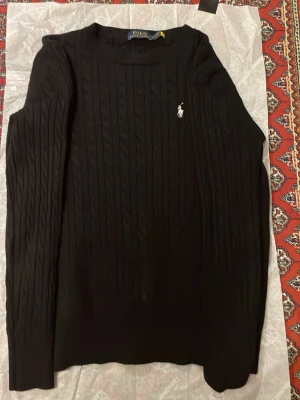 Svart kabelstickad tröja Polo Ralph Lauren - Kabelstickad svart tröja från Polo Ralph Lauren med klassisk vit logga på bröstet. Tröjan har rund halsringning och långa ärmar. Perfekt för dig som gillar stilrena och tidlösa plagg. Materialet är väldigt mjukt. Köpte även denna på Corner i Mallorca, såklart äkta! Vid minsta fråga hör av dig till mig😁😊! St:M men den passar S också.
