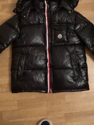 Svart Moncler pufferjacka L - Säljer en svart pufferjacka från Moncler i storlek L. Jackan har huva, två sidofickor, en bröstficka med Moncler-logga och dragkedja med rödvitblå detalj. Materialet är glansigt och fyllt med dun, perfekt för kalla dagar.