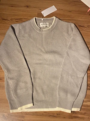 Grå ribbad stickad tröja från Maison Margiela - Säljer en stilren grå stickad tröja från Maison Margiela med ribbad struktur och ljusa detaljer vid muddar och nederkant. Tröjan har rund hals och långa ärmar, perfekt för lager-på-lager under kyliga dagar.
