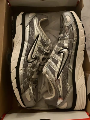 Nike P6000 - Säljer ett par Nike P6000 i silver och grått med vita detaljer. Skorna har ett futuristiskt mönster med mesh och blanka paneler, samt en stor svart Swoosh på sidan. Klassisk snörning och tjock vit sula med svart yttersula för extra grepp.