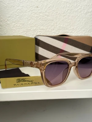 Burberry solglasögon i beige plast - Snygga Burberry solglasögon med transparent beige båge i plast och lila tonade glas. Sidorna har ett rutigt mönster och en metallogo med B. Kommer med originalfodral, putsduk och box.