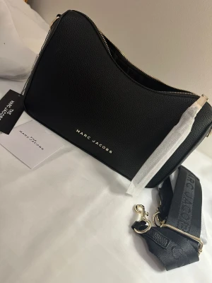 Svart axelväska  - Snygg svart axelväska från  i strukturerat skinn med guldfärgade detaljer och dragkedja upptill. Väskan har en bred, justerbar axelrem med logotyp och stilren form. Perfekt för dig som vill ha en trendig och praktisk väska med exklusiv känsla.