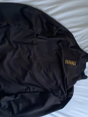 Svart track kofta från EA7 Emporio Armani - Säljer en svart track kofta från EA7 Emporio Armani med guldfärgade detaljer och logga på bröstet. Jackan har dragkedja framtill, hög krage och är tillverkad i ett mjukt syntetmaterial. Perfekt för en sportig och clean look. Perfekt inför vår och höst.