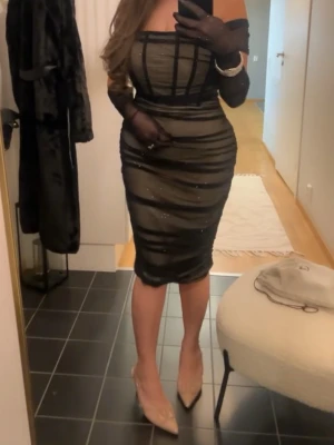 Svart glittrig meshklänning - Supersnygg svart kortklänning i mesh med glittriga detaljer och transparenta partier. Klänningen har figurnära passform, offshoulder och rynkade sömmar som ger en cool look. Perfekt för dig som vill sticka ut med en trendig och elegant stil.Passar en S-M i storlek medföljer matchande handskar.